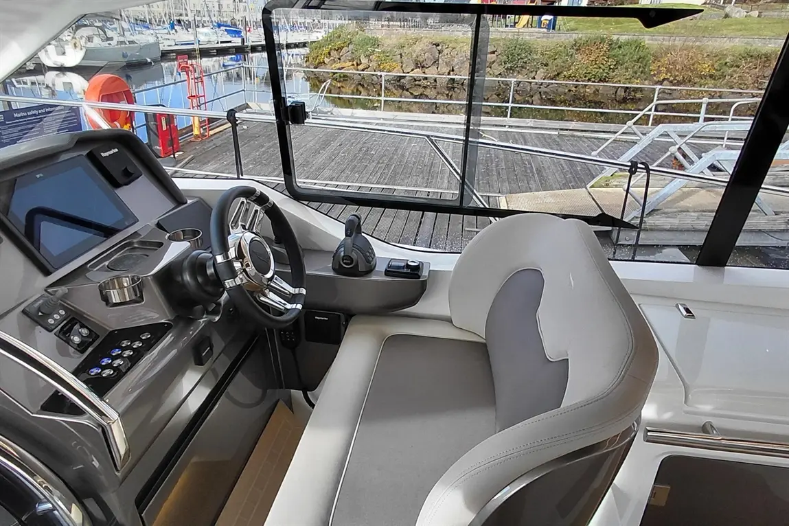 Boat cockpit view featuring a steering wheel, control panel, and comfortable seating, with a scenic marina backdrop.